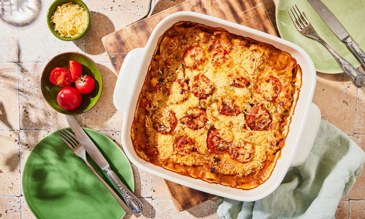 Een witte ovenschaal met een goudbruine lasagne, gegarneerd met gesmolten kaas en plakjes tomaat, staat op een houten plank. Naast de schaal liggen twee groene borden met bestek, een kleine groene kom met geraspte kaas en een andere met verse tomaten, alles op een lichtgekleurde tegelvloer met een groene doek erbij.