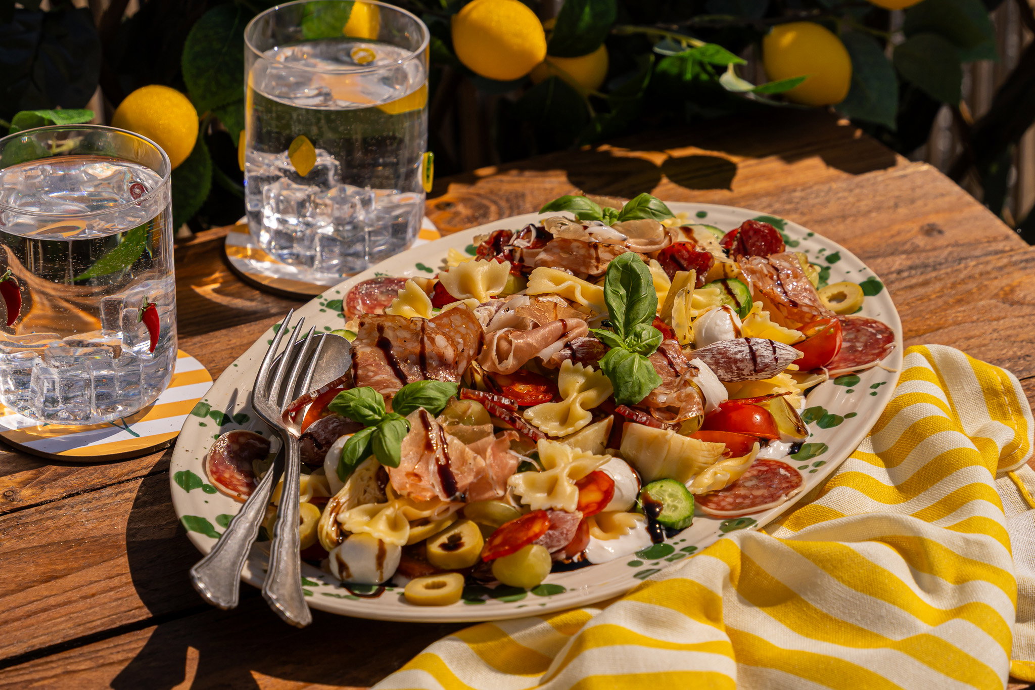 Kleurrijke antipasti-pastasalade met farfalle, burrata, cherrytomaten, komkommer, druiven, olijven, artisjokken en verschillende Italiaanse vleeswaren, afgewerkt met verse basilicum en balsamicocrème.