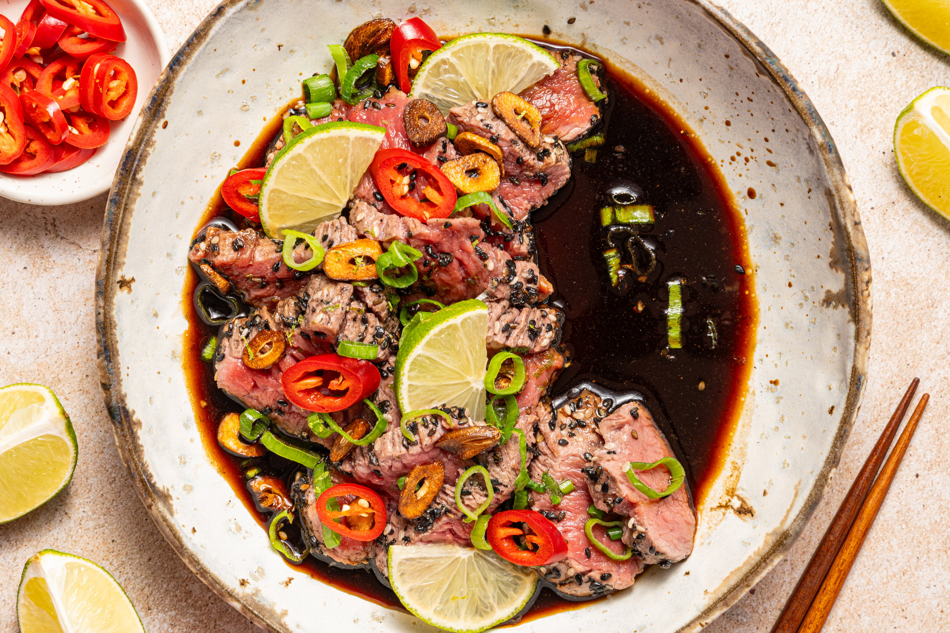 Beef tataki met crispy knoflook en yuzu ponzu in een kom met limoen en rode peper.