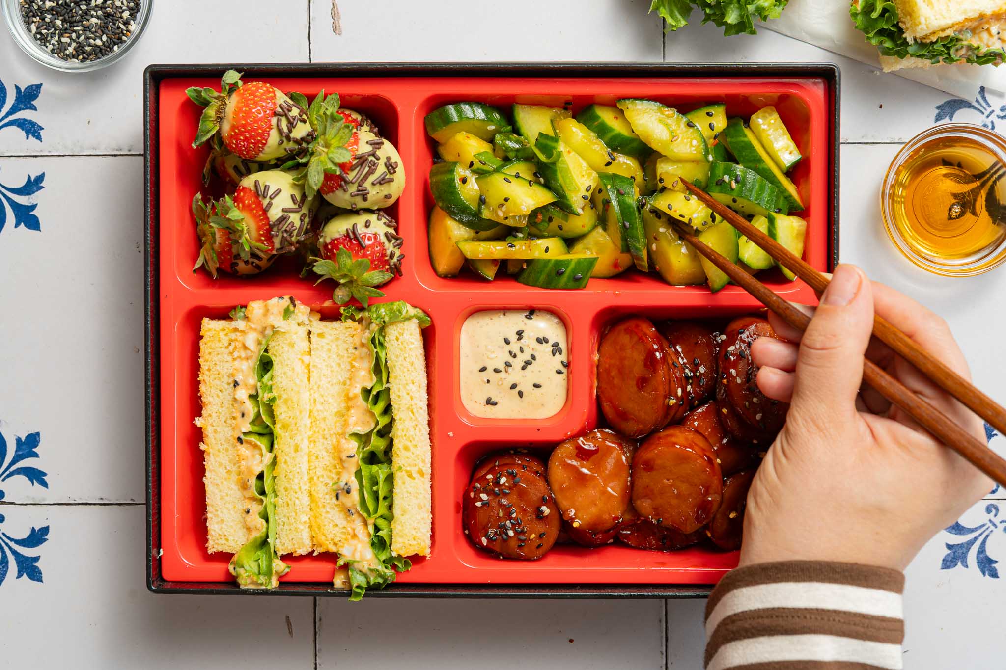 Aziatisch-Nederlandse fusion bento box met teriyaki rookworst, gochujang eiersalade sandwiches, matcha aardbeien en piccalilly komkommer.